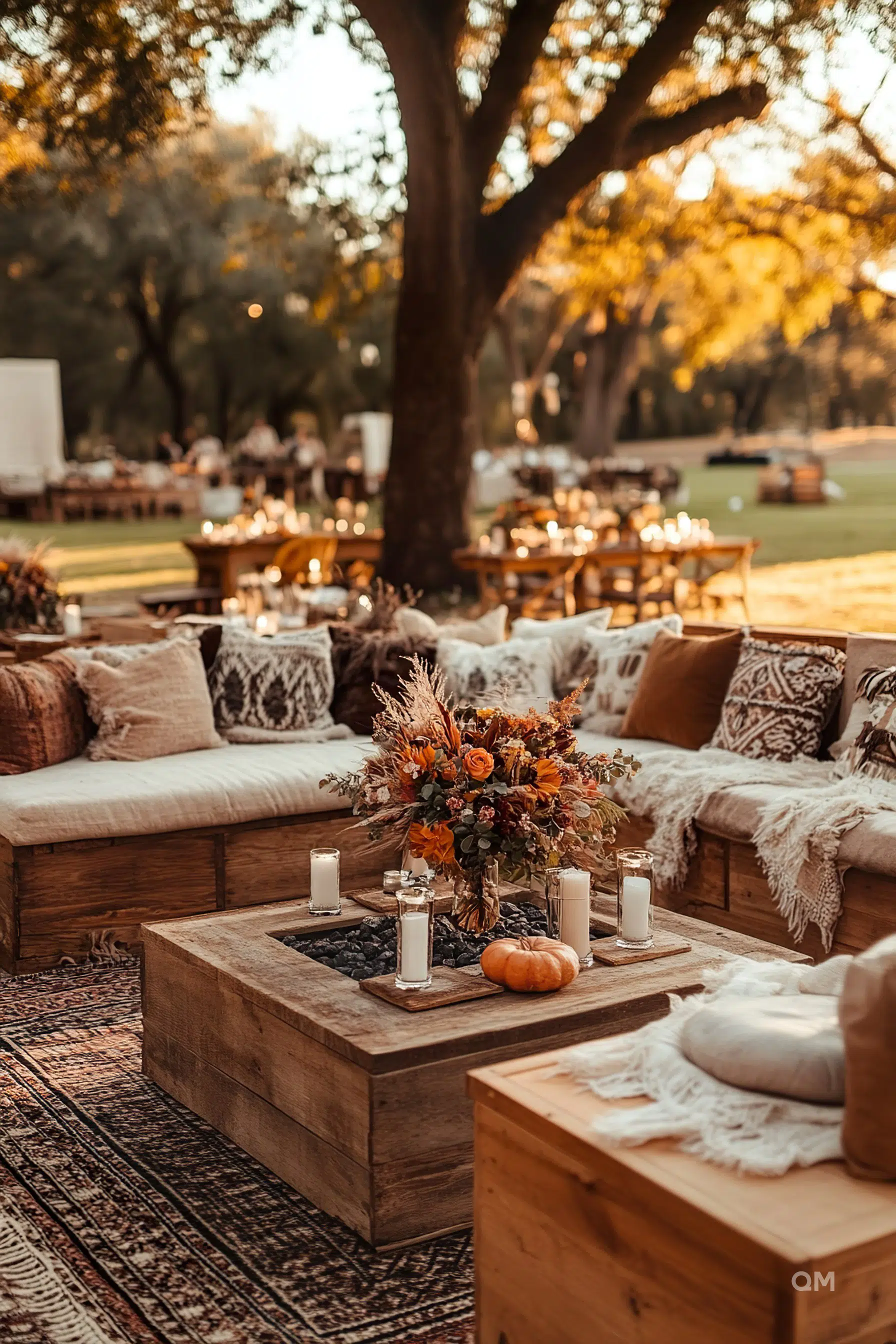 Outdoor autumnal event setting with wooden benches, a floral centerpiece, candles, and a pumpkin on a rug under trees.