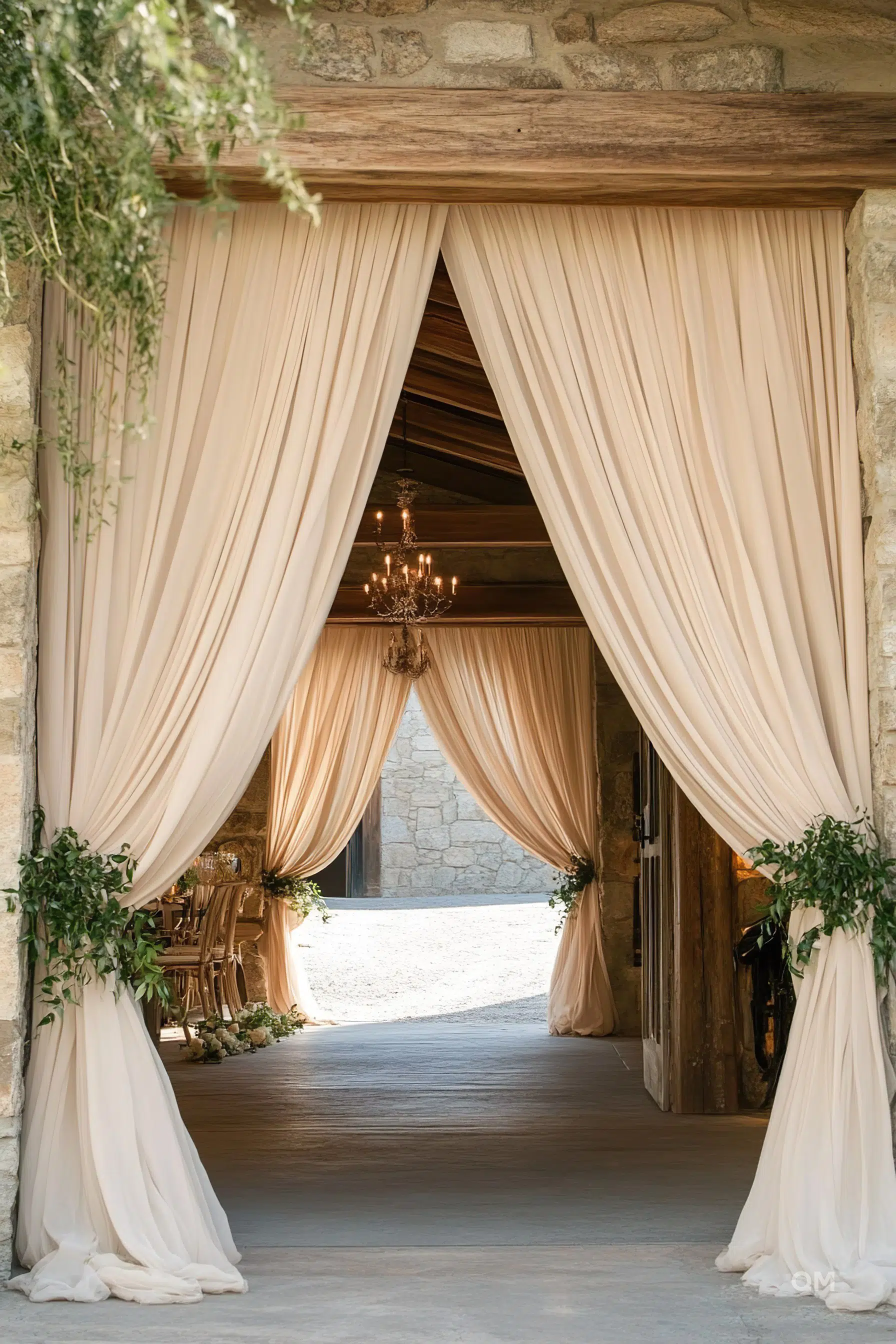 Elegant beige drapes frame the entrance to a rustic stone building with a visible chandelier inside and greenery adorning the sides.