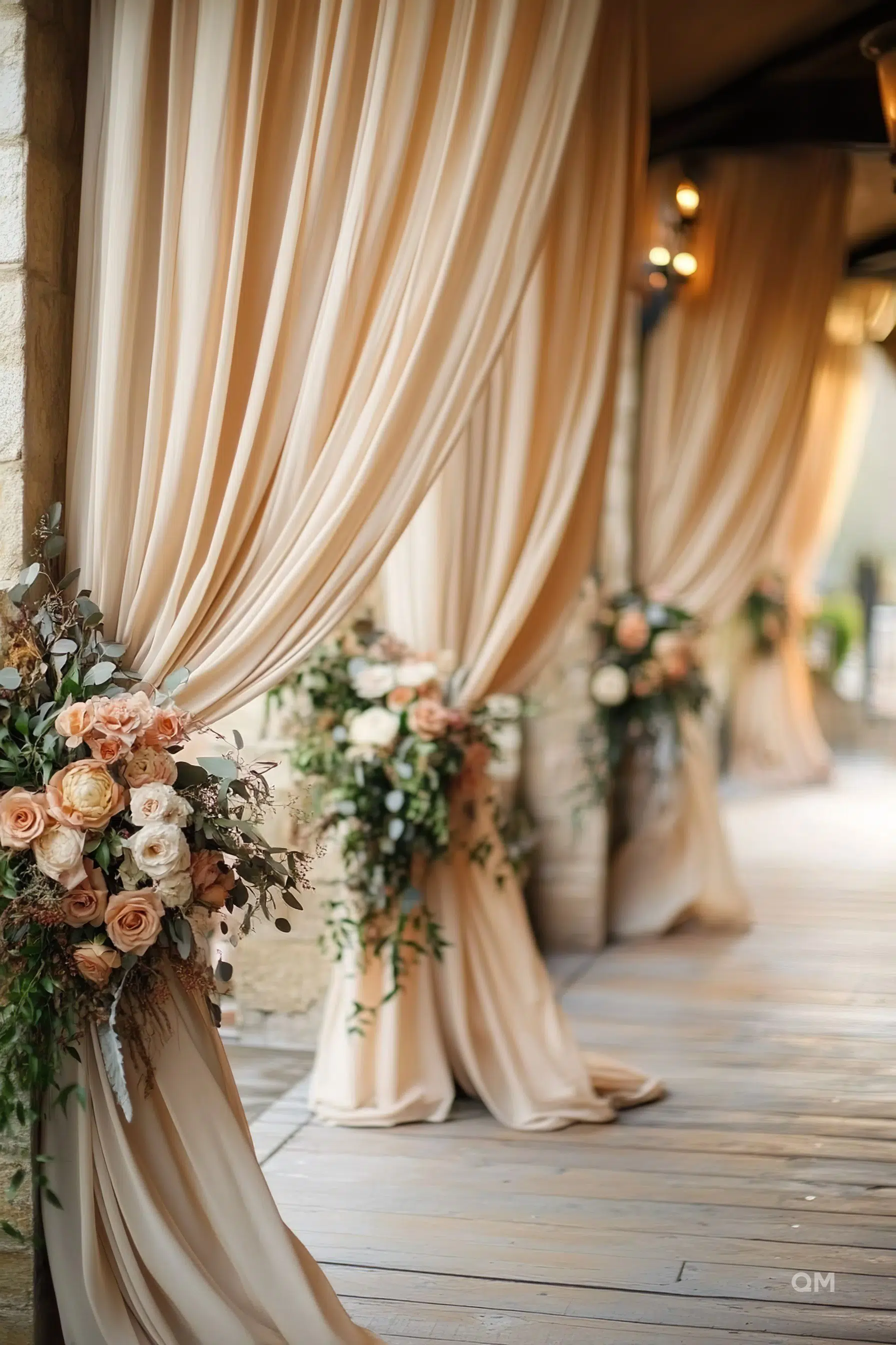 Elegant beige draperies tied with floral arrangements on a stone wall, leading to a wooden floor pathway.