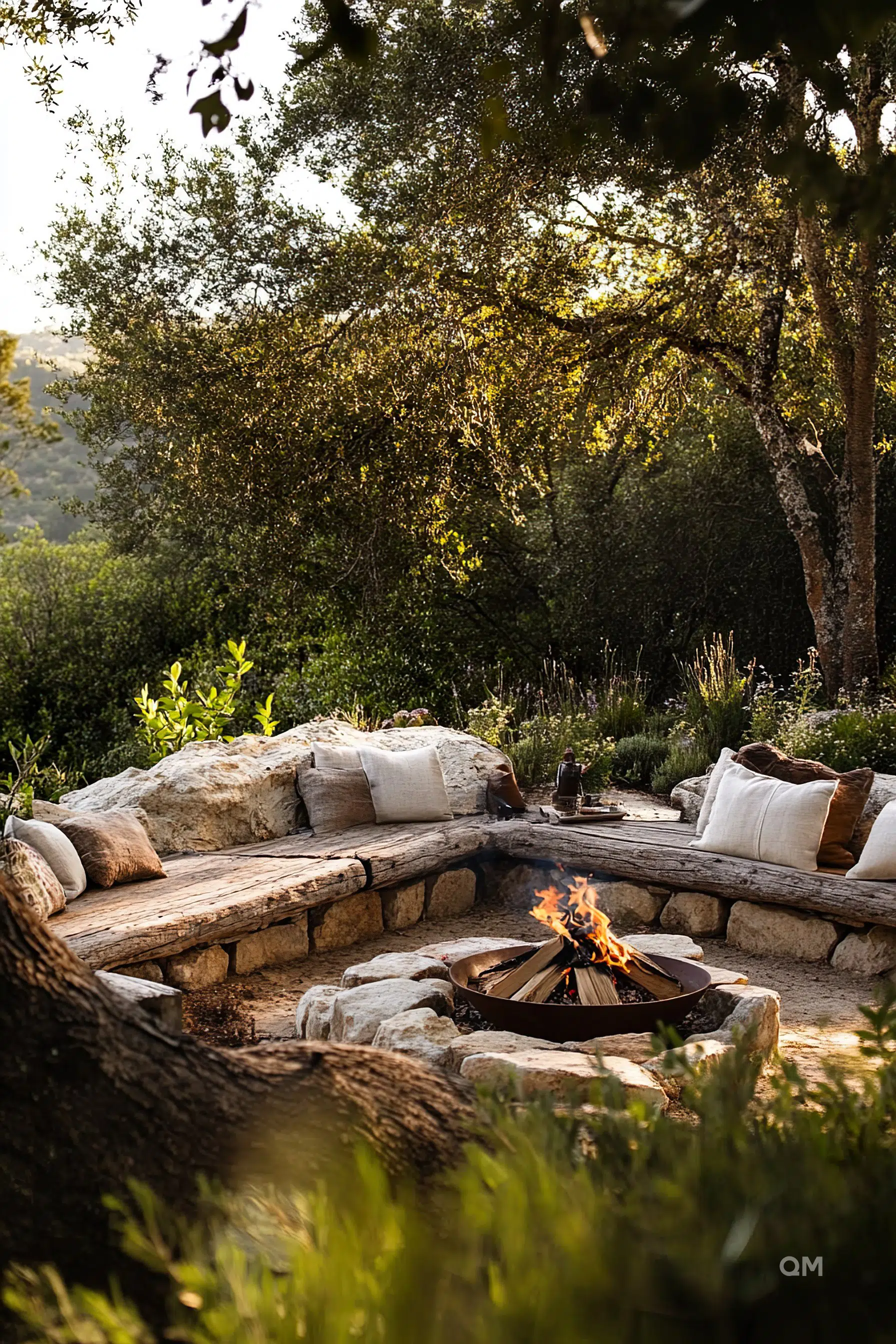 Rustic outdoor fire pit with benches adorned with cushions amid trees and greenery at dusk.