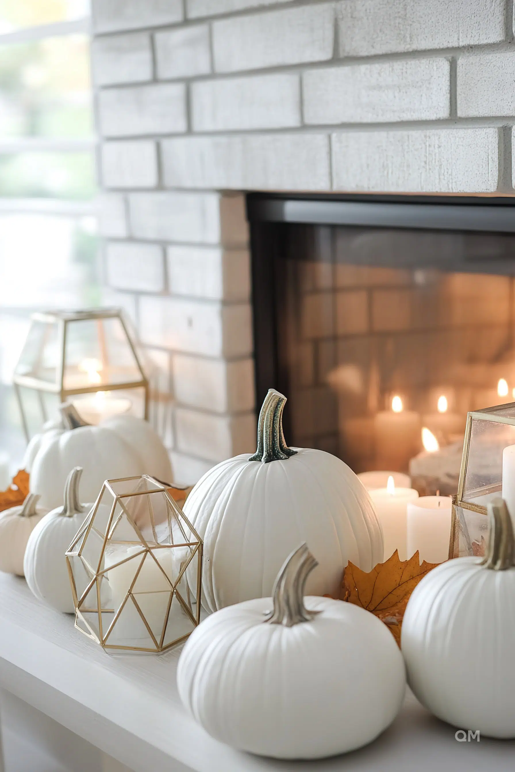 Decorative setting with white pumpkins, lit candles, and geometric candle holders on a mantlepiece.