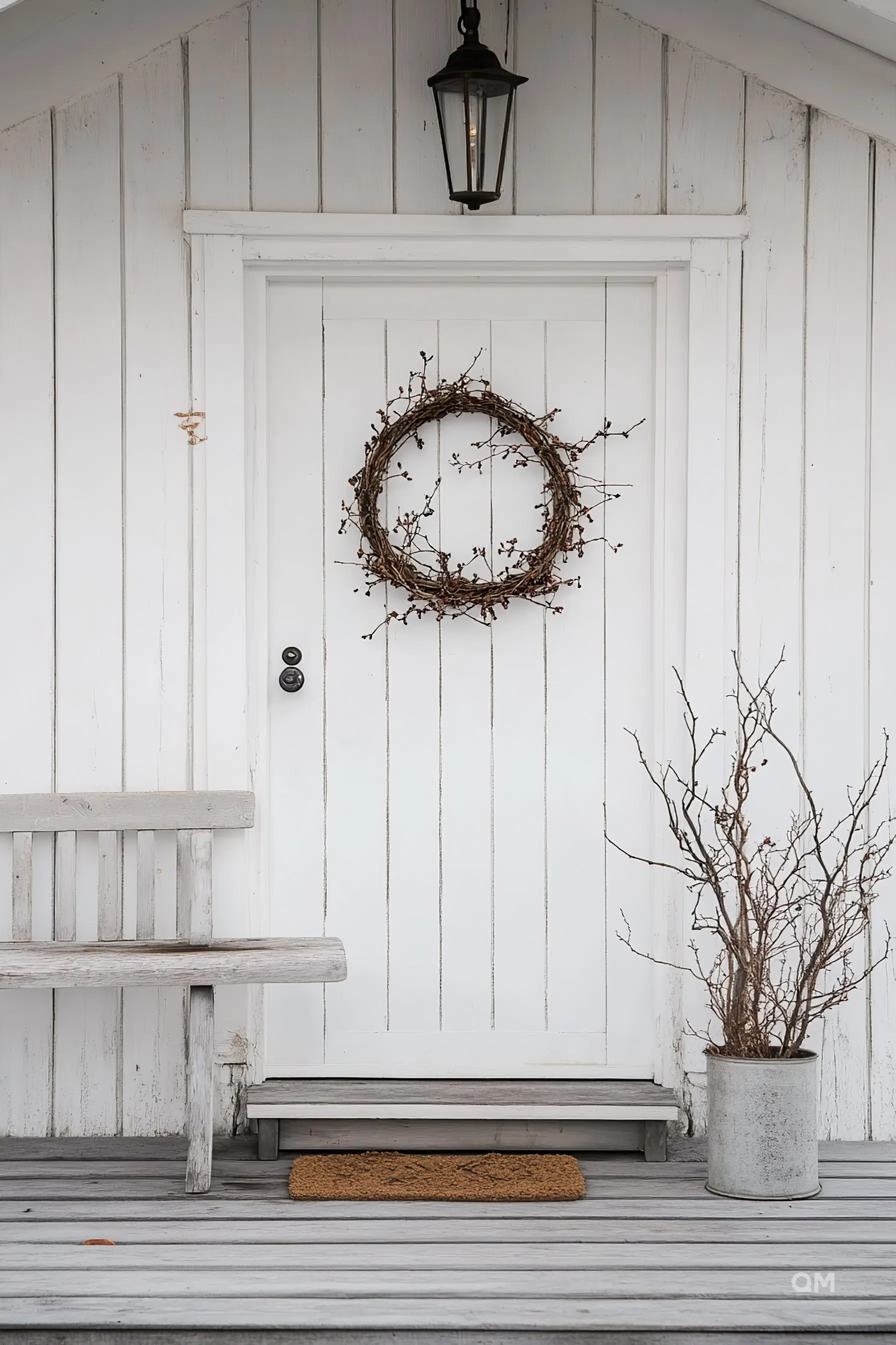 A simple white door with a twig wreath, outdoor lantern, wooden bench, doormat, and potted dried branches on a porch.