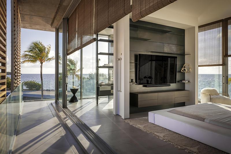 primary bedroom with full-height sliding glass wall open to waterfront terrace and palm tree view, low white platform be