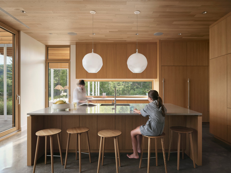 kitchen with flat-front oak cabinetry, grey stone island, round oak stools, two white globe pendants, horizontal window