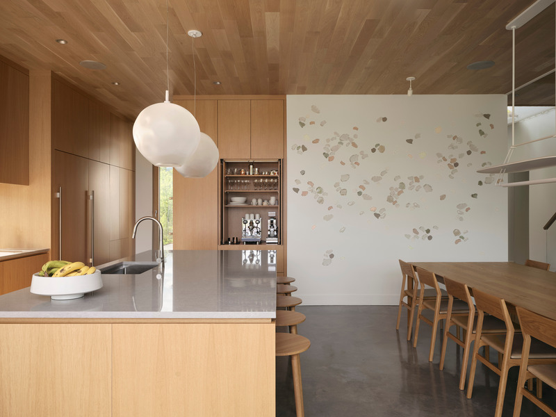 kitchen island with grey stone countertop and oak cabinetry transitioning to dining area, white plaster wall with scatte