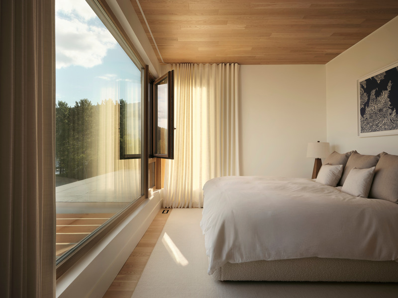 primary bedroom with floor-to-ceiling oak-framed window open to lake view, ivory linen drapes in warm light, low platfor