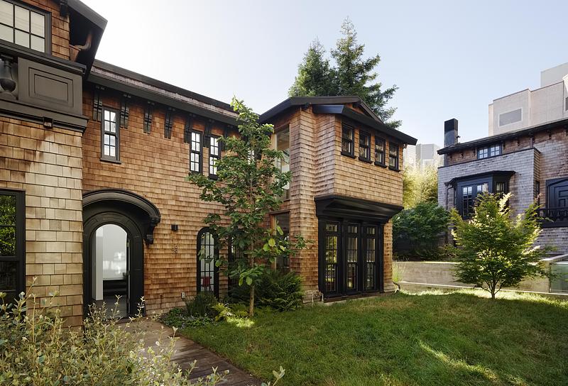 rear garden elevation of two-story cedar shingle house with black-framed arched entry door, young trees on green lawn, a