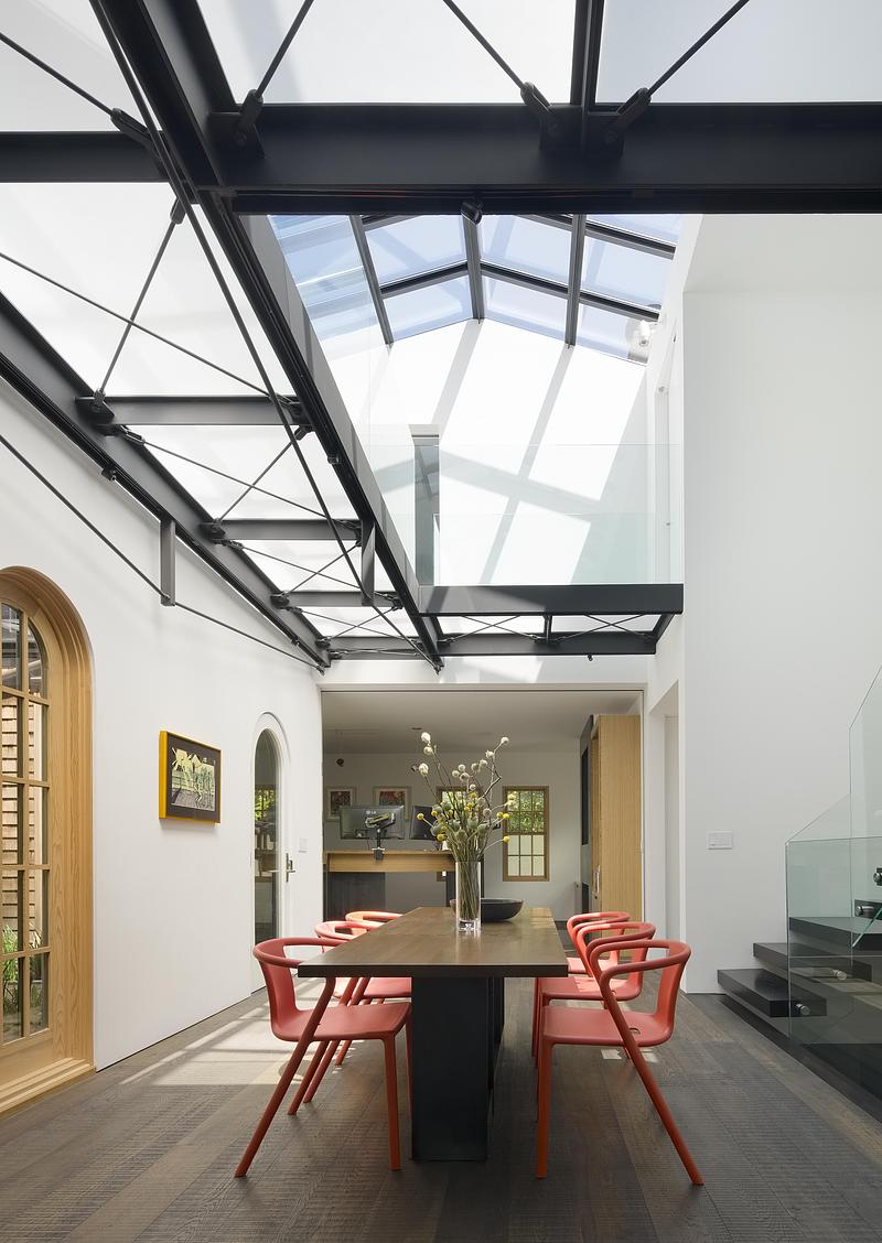 interior dining room under black steel truss glass roof, long dark wood table with coral-red armchairs, white walls, dar