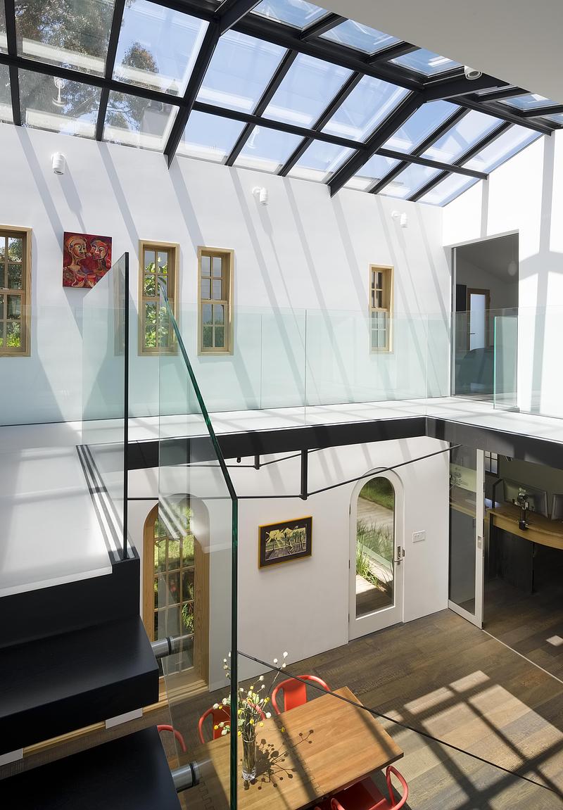 double-height interior showing black steel floating stair with glass balustrade, coral dining chairs in foreground, arch