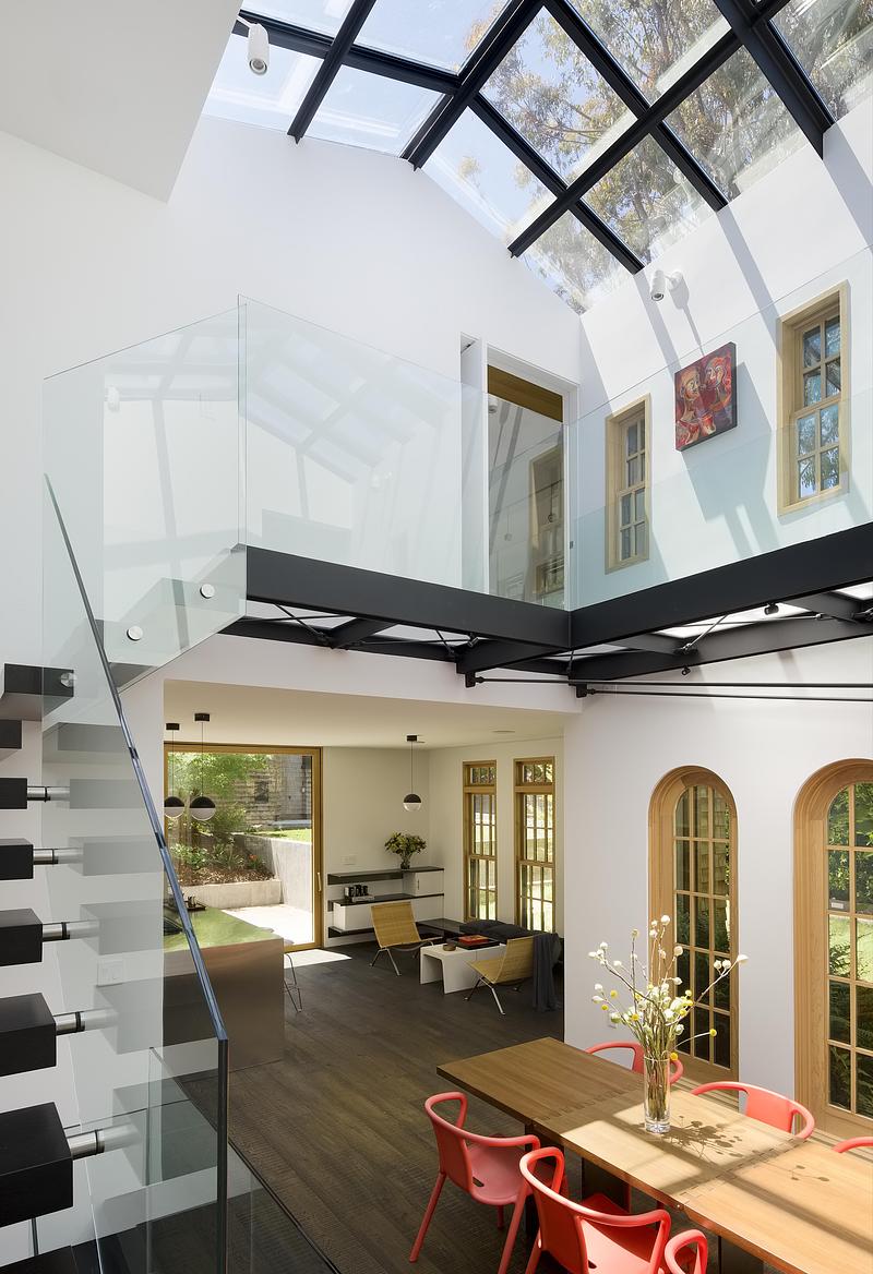 interior dining room angle showing full glass roof with black steel trusses, wood dining table with coral chairs, glass