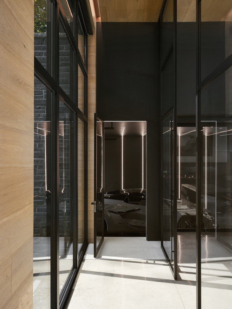 glass-walled corridor with black steel-framed pivot doors opening to a dark garage lit by vertical linear strips, oak-pa