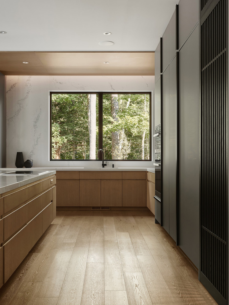 kitchen perimeter wall showing oak lower cabinets with white stone countertop, black-framed window looking into dense gr