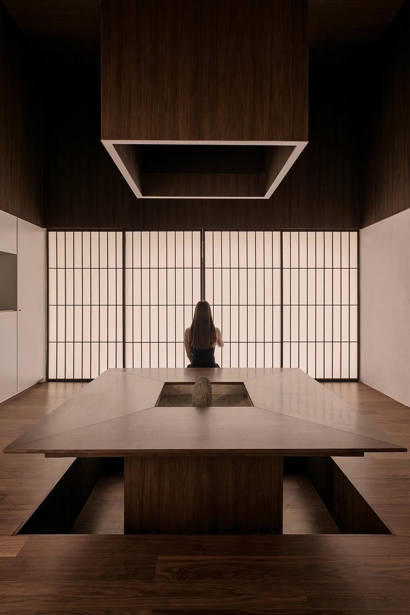 double-height dining room with dark walnut table and floor, figure seated facing full-height glowing shoji rice-paper sc