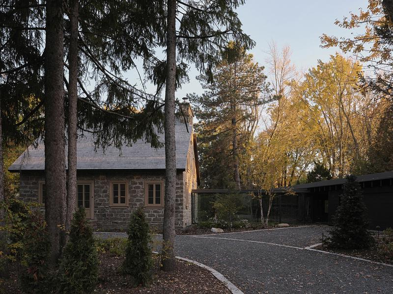 exterior ground-level view of fieldstone house facade at dusk with warm-lit French doors, slate roof, tall pines, and bl