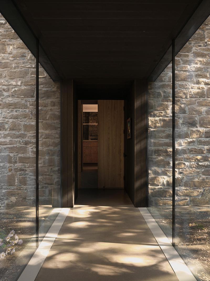 entry passage through glass connector with exposed fieldstone walls on both sides, dark steel ceiling, dappled tree shad