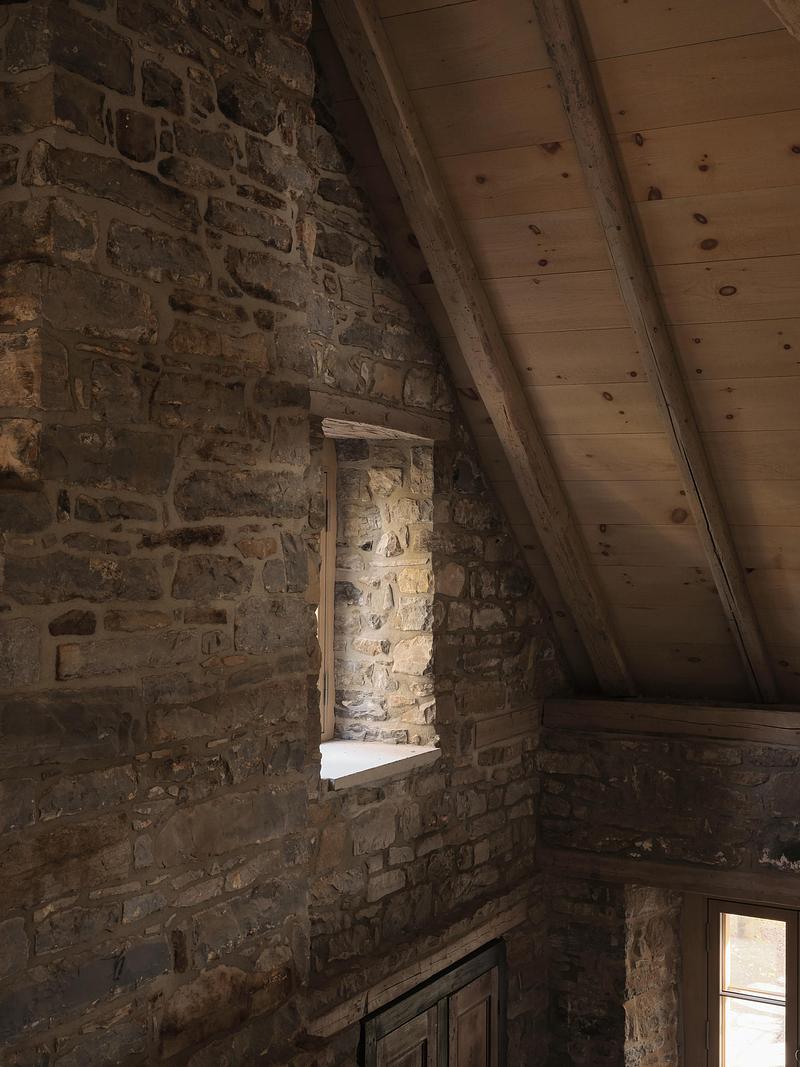 upper stair landing view of rough rubble fieldstone wall with single deep-set window opening, steep knotty pine ceiling