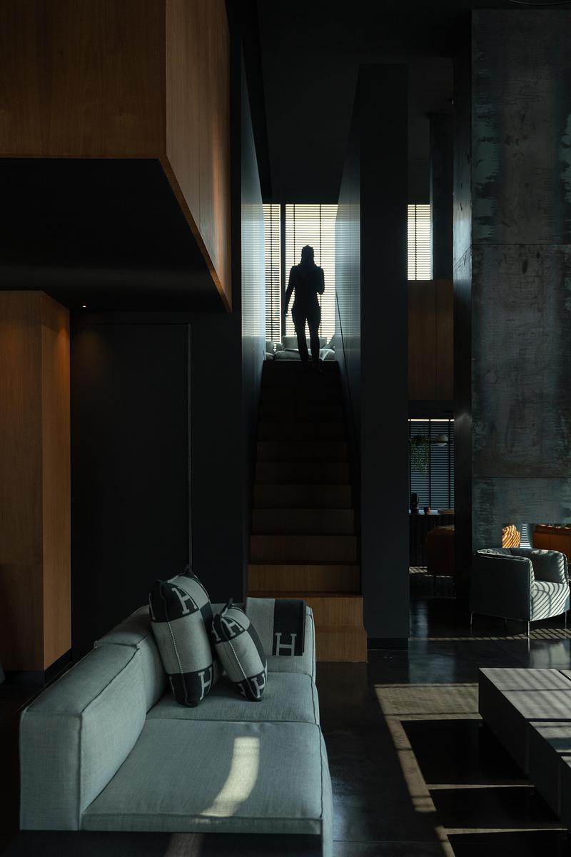 double-height staircase hall with silhouetted figure at top, pale linen sofa with monogram cushions below, warm oak mill