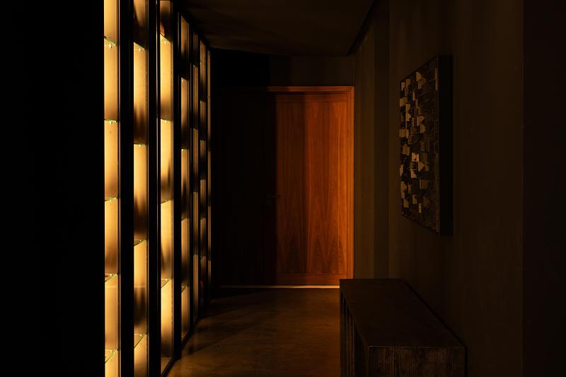 private corridor with amber-backlit glass display cabinet lining left wall, full-height warm-grain timber door at end, g