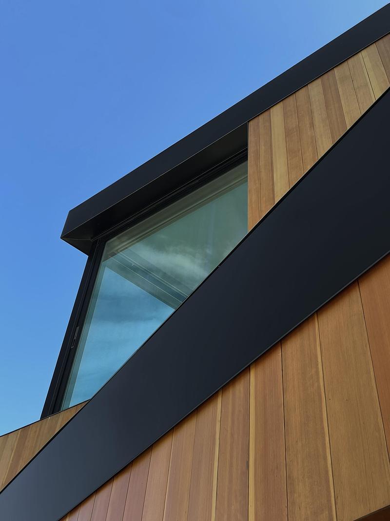 close-up exterior detail of upper corner of cedar-clad house showing angled matte black fascia framing a large corner gl
