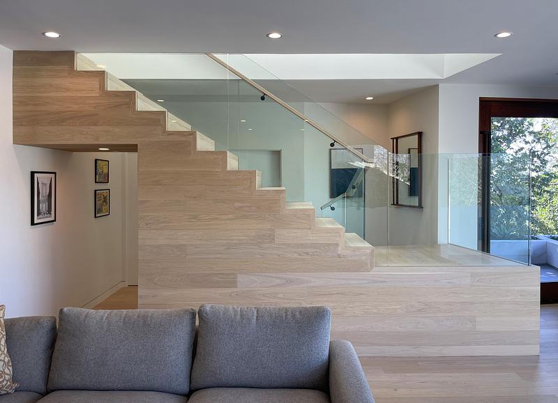 living room view toward pale white oak stair with glass balustrade, framed artwork hung on white wall under stair soffit