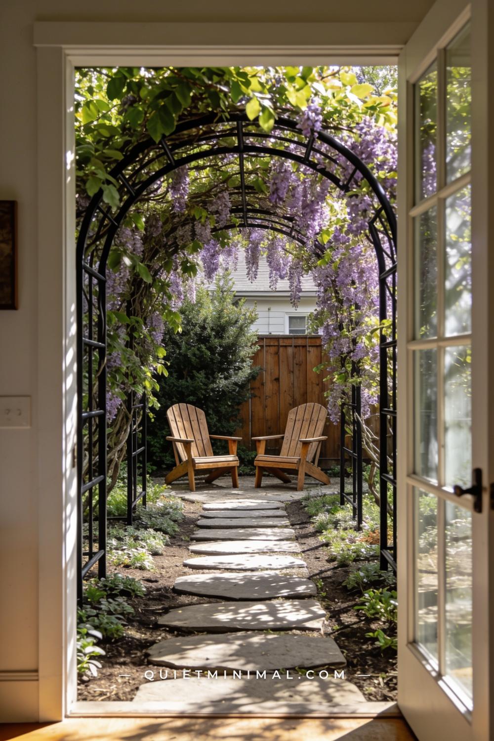 A vertical photograph captures a wide garden path where a simple metal arch — raw or powder-coated black steel — support