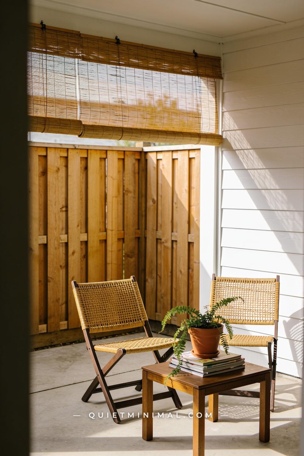 A vertical photograph captures a sunny patio corner where a bamboo roll-up privacy screen is mounted along the top of a