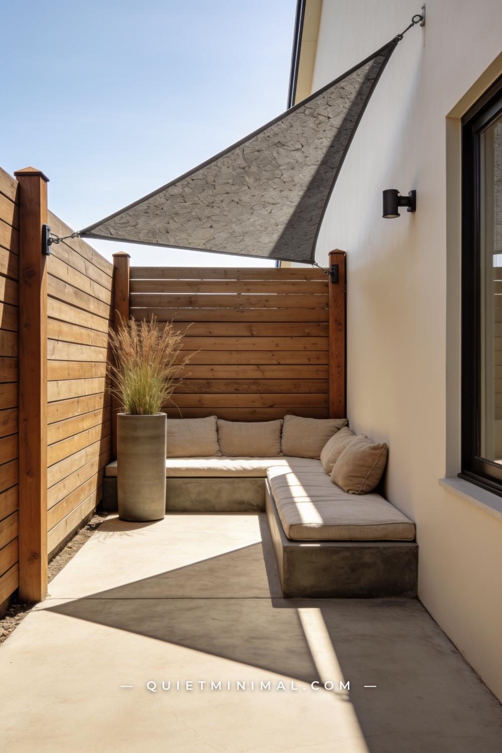 A vertical photograph captures a small square concrete patio with a triangular shade sail in warm stone gray stretched d