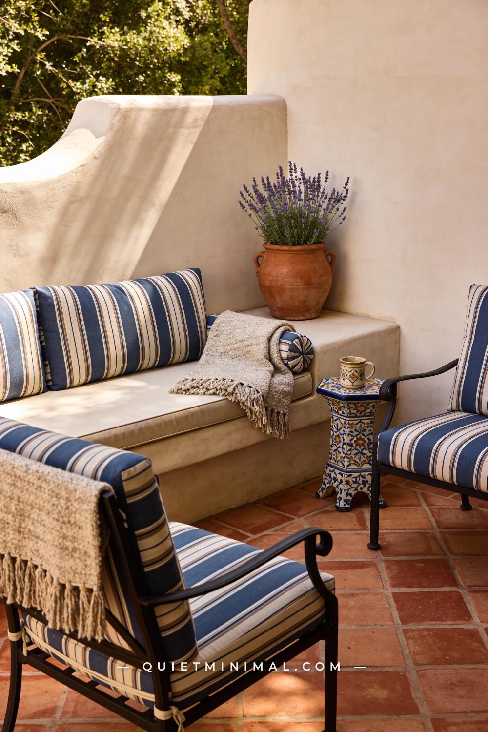 A wide photograph of a Spanish Revival patio lounge corner featuring a low bench and two chairs draped with striped outd
