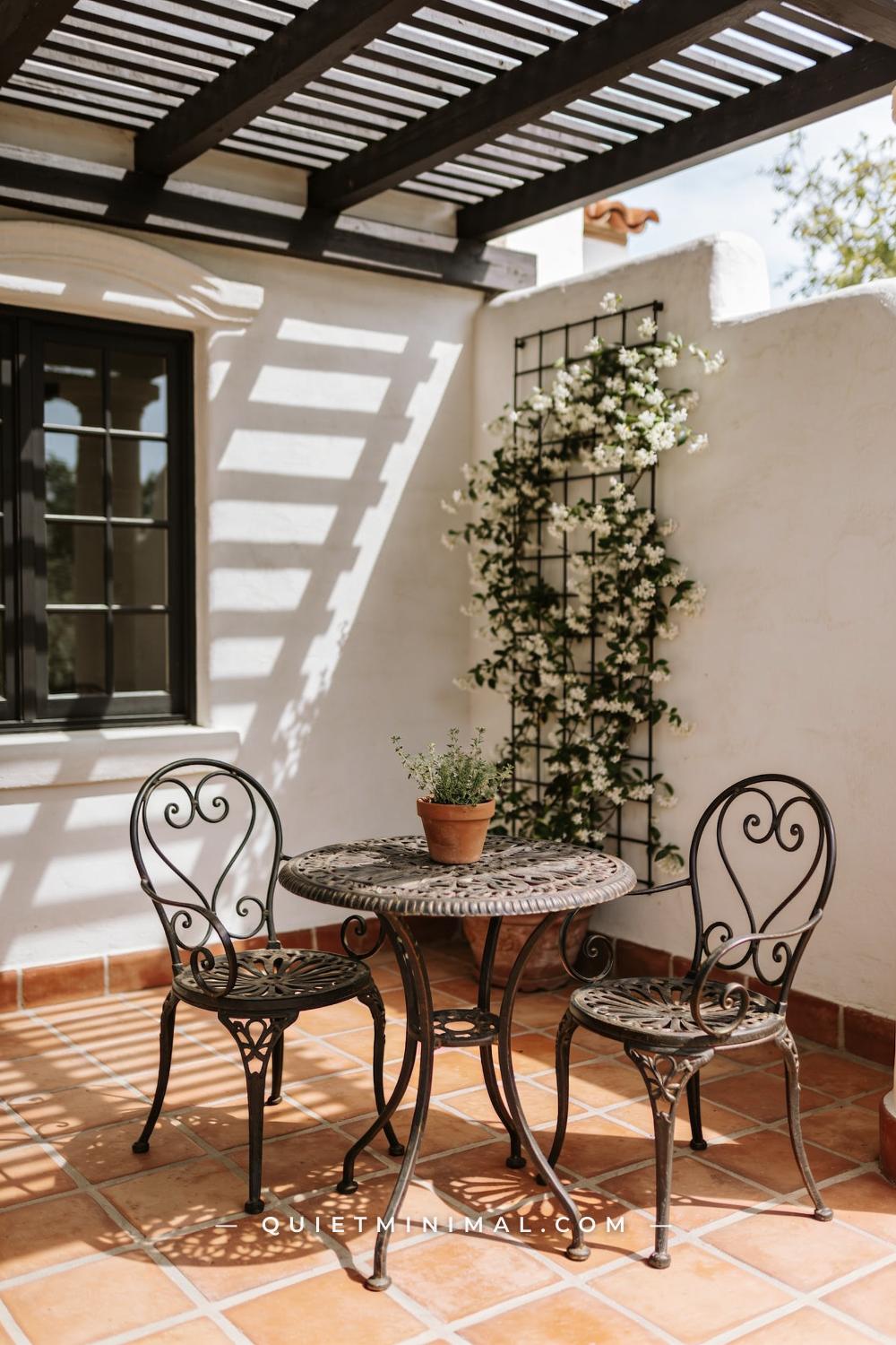 A medium-wide photograph of a Spanish Revival patio seating area centered on a wrought iron bistro table and two matchin
