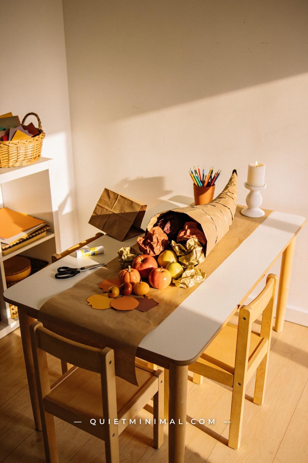 A wide establishing shot from a straight-on wall view of a kids table showing a central cornucopia craft in progress: br